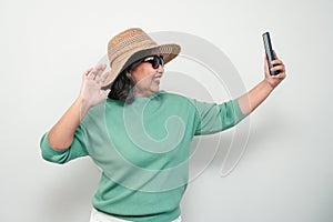 Portrait of happy Asian senior woman wearing sunglasses, a hat, and holding smartphone for a selfie, standing isolated on white