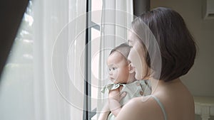 Portrait of happy asian woman mother holding baby infant near window looking away at home. 4 months baby embracing in parent arm.