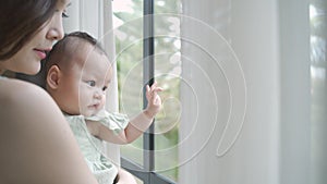 Portrait of happy asian woman mother holding baby infant near window looking away at home. 4 months baby embracing in parent arm.
