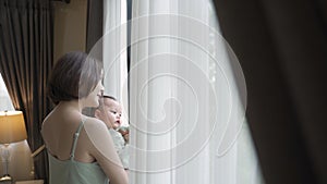 Portrait of happy asian woman mother holding baby infant near window looking away at home. 4 months baby embracing in parent arm.