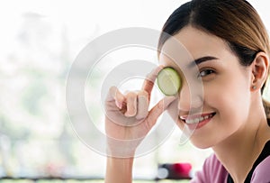 Portrait of happy Asian woman professional masseuse in uniform standing holding a sliced cucumber and paste at the eye in spa