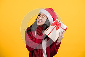 Portrait of a happy Asian woman wearing a warm sweater in a Christmas hat standing alone on a yellow background holding a gift box