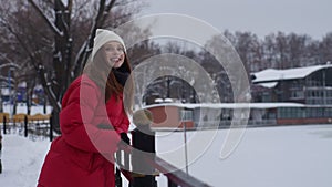 Portrait of happy attractive young woman in hat and red fashion warm jacket standing in snowy city park and cheerfully