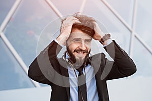 Portrait of happy bearded businessman with headphones on neck, skycraper windows background.
