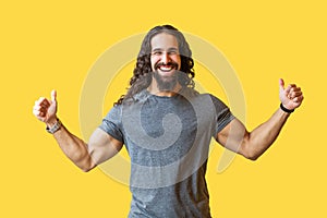 Portrait of happy bearded young man with long curly hair in grey tshirt standing with thumbs up and looking at camera with toothy