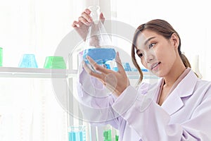 Portrait of happy beautiful Asian scientist woman in lab coats holding and looking at blue chemical flasks. Researcher doing