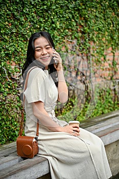 A happy Asian woman is on the phone with her friend while sitting on a bench in the city