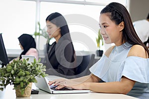 Portrait of happy beautiful Asian woman officer sitting at office desk, typing on laptop computer with blurred background of busy