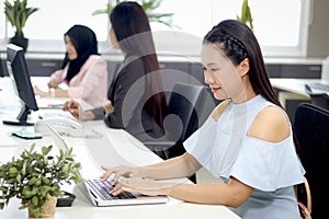 Portrait of happy beautiful Asian woman officer sitting at office desk, typing on laptop computer with blurred background of busy
