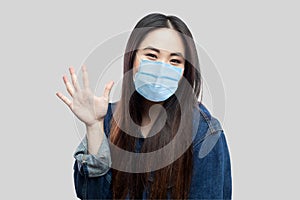 Portrait of happy beautiful asian young woman with surgical medical mask in denim jacket standing and waving her hand and greeting