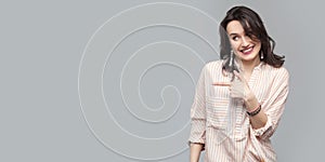 Portrait of happy beautiful attractive brunette woman in casual striped orange shirt and curly hairstyle standing and pointing at