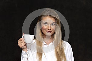 Portrait of a happy and beautiful blonde 20-25 years old with a white cup in a long shirt on a black textured background.