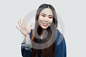Portrait of happy beautiful brunette asian young woman in blue denim jacket, makeup standing and waving her hand and greeting