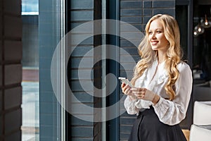 Portrait of happy beautiful businesswoman holding mobile phone, smiling and looking sideways. standing near window, indoor