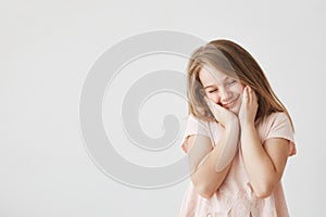 Portrait of happy beautiful girl with blond hair in pink t-shirt squeezing face with hands, closing eyes, feeling shy