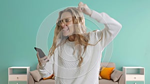 Portrait of a happy beautiful girl in glasses is looking to a mobile phone and celebrating at blue room background