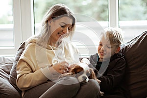 Portrait of happy beautiful middle-aged woman sitting on grey sofa with teenage boy at home, holding dog, stroking pet.