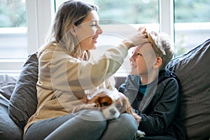 Portrait of happy beautiful middle-aged woman sitting on sofa with teenage boy at home, holding dog, patting sons head.