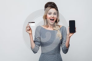Portrait of happy beautiful modern young girl in grey dress standing, holding cell phone and credit card with open mouth, looking
