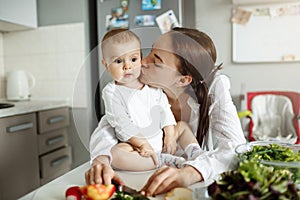 Portrait of happy beautiful mother kissing her lovely baby in cheek in dinner room. Baby sitting on table with surprised
