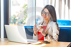 Portrait of happy beautiful stylish brunette young woman in glasses sitting, resting, enjoying, holding cup of coffee and looking