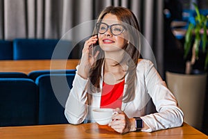 Portrait of happy beautiful stylish brunette young woman in glasses sitting talking on mobile smart phone and toothy smile