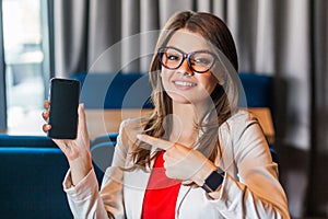 Portrait of happy beautiful stylish young woman in glasses sitting, showing and pointing at mobile smart phone screen and looking