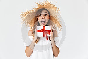 Portrait of happy beautiful woman 20s wearing big straw hat smiling and holding present box with red ribbon, isolated over white