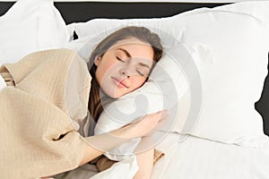 Portrait of happy, beautiful young woman resting in bed, wearing pyjamas, hugging her pillow and smiling while sleeping