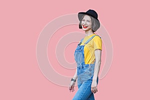 Portrait of happy beautiful young woman in yellow t-shirt and blue denim overalls with makeup and black hat standing and looking