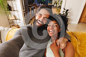 Portrait of happy biracial bearded man holding girlfriend's hands and taking selfie on sofa