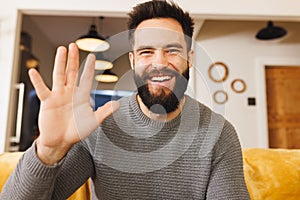 Portrait of happy biracial bearded young man waving hand while sitting on sofa in living room