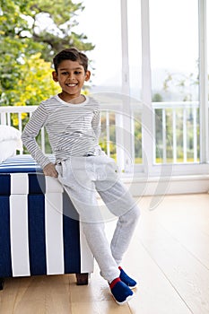 Portrait of happy biracial boy wearing pyjamas in bedroom in front of window with view of garden