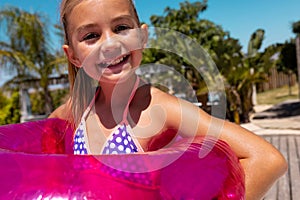 Portrait of happy biracial girl smiling and holding swim ring by the swimming pool