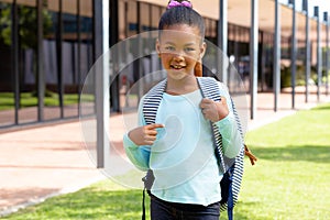 Portrait of happy biracial schoolgirl with school bag smiling in schoolyard