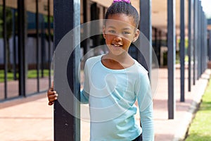 Portrait of happy biracial schoolgirl smiling, holding post in sunny schoolyard