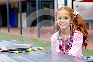 Portrait of happy biracial schoolgirl at table with books and tablet in schoolyard