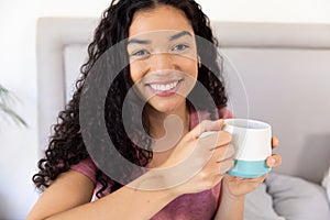 Portrait of happy biracial woman holding cup of coffee sitting on bed at sunny home