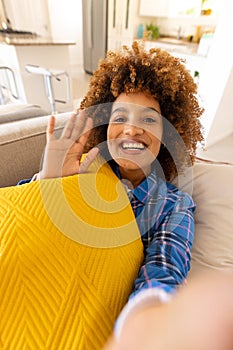 Portrait of happy biracial young woman with afro hair waving hand while resting with cushion on sofa