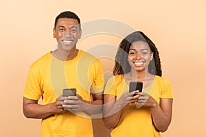 Portrait of happy black couple using cellphones and smiling at camera, standing on yellow studio background