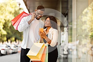 Portrait of happy black couple using phone with shopping bags