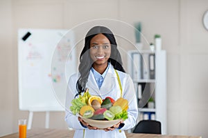 Portrait of happy black dietitian in lab coat holding bowl of fresh fruits and vegetables, smiling at camera at clinic