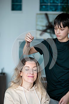 Portrait of happy blond middle-aged woman sitting with closed eyes getting head massage from teenage boy using massager.