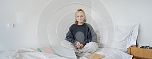 Portrait of happy blond woman, freelancer working from home, sitting on bed with laptop and notebooks. Student doing