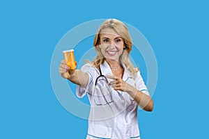 Portrait of happy blonde caucasian mature female doctor pointing at orange bottle of pills.