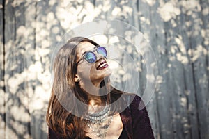 Portrait of happy boho girl in cool outfit and sunglasses smiling in sunlight. Stylish hipster girl posing in sunny street on