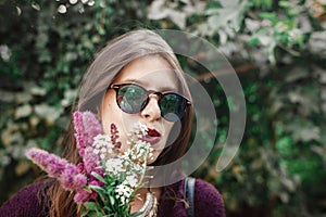 Portrait of happy boho girl in sunglasses smiling with bouquet of wildflowers in sunny garden. Stylish hipster carefree girl