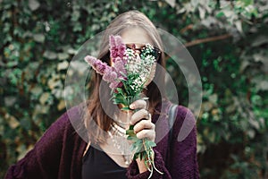 Portrait of happy boho girl in sunglasses smiling with bouquet of wildflowers in sunny garden. Stylish hipster carefree girl