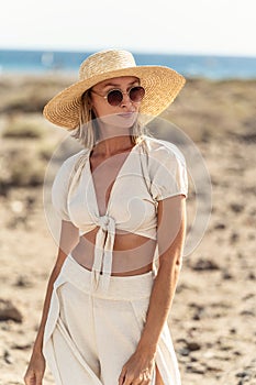 Portrait of happy boho woman wearing straw hat, sunglasses and fashionable summer clothes, posing on the wild beach