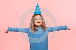 Portrait of happy brunette little girl with funny party cone on head raising hands in happiness
